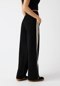 Schwarze weit geschnittene Hosen aus weichem Stoff, mit vertikalen elfenbeinfarbenen Streifen an den Seiten und einem elastischen Bund.