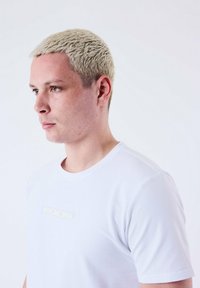 Camiseta blanca de cuello redondo hecha de tela suave, presenta un sutil logo PXP en un color claro en la parte frontal, mangas cortas, diseño ajustado.
