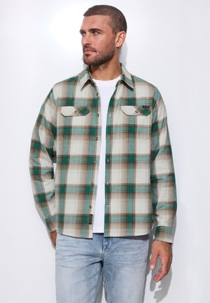 Mann trägt ein grün-braun kariertes Hemd über einem weißen T-Shirt und hellblauen Jeans, steht vor weißem Hintergrund und schaut nach links.