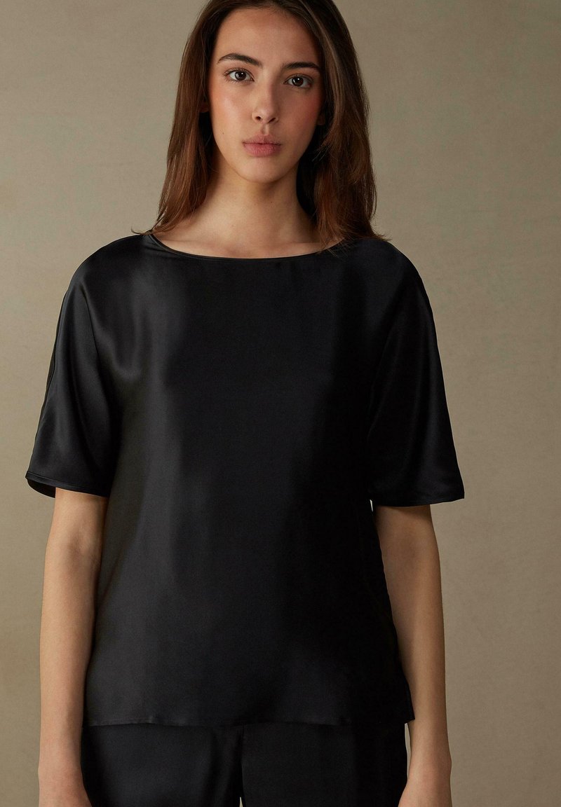 Top de seda negra de manga corta con escote barco. Textura suave, corte holgado y detalles de costura mínimos en los bordes.
