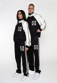 Sorte træningsdragter med hvide accenter, der har et grafisk logo på forsiden. Outfits har ribbede manchetter og striber ned ad bukserne.