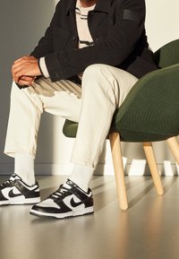 Man sitter på en grön stol klädd i svart jacka, beige byxor, vita strumpor och svart-vita Nike-sneakers inomhus.