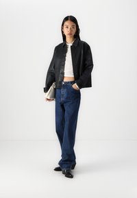 Must last nahk cropped jakki nupuga, koos kõrge vöökohaga siniste denim pükstega. Mudel hoiab heledat käekotti.