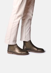 Bronzefarbene metallische Chelsea-Stiefel mit elastischen Seitenteilen, flachen Sohlen und einer glatten Textur, getragen mit off-white Jeans mit ausgefranstem Saum.
