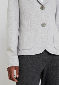 Šedé strukturované sako s klopovým límcem, dvěma předními kapsami a třemi kulatými knoflíky. Kombinováno s tmavými kalhotami.