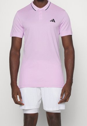 Homme portant un polo à manches courtes violet clair avec un logo noir et un short blanc superposé, debout devant un fond uni.