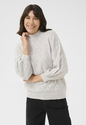 Pull en tricot gris clair avec un col montant, un motif texturé et des manches bouffantes, associé à un jean foncé. Design doux et décontracté.