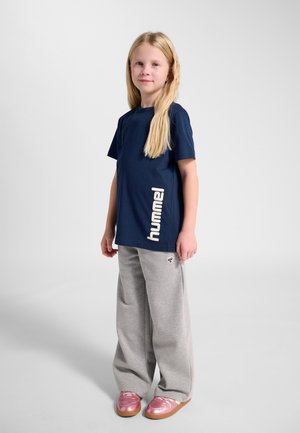 Blondynka stoi przodem, ubrana w granatową koszulkę hummel, jasnoszare szerokie spodnie i metalicznie różowe buty na jednolitym tle.