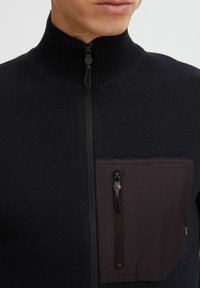 Svart zip-up genser med ribbekrage. Har en mørk stofflomme foran med glidelås, teksturert strikkemateriale og kontrasterende stoffpanel.