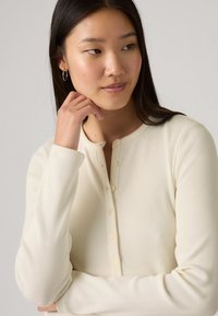 Μπλούζα μακρυμάνικη, κρεμ χρώματος με ραβδώσεις, που διαθέτει στρογγυλή λαιμόκοψη και πέντε κουμπιά στη ραφή. Το ύφασμα είναι ομαλό με ελαφριά ελαστικότητα.