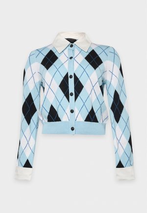 Une femme porte un cardigan bleu clair à motifs losanges, un pantalon noir à jambes larges et des chaussures plates marron foncé, sur un fond de studio blanc uni.