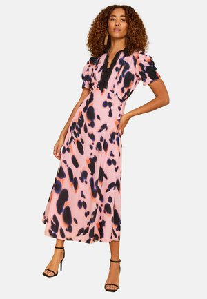 Frau mit lockigem Haar, die ein pinkes Midikleid mit schwarzen und violetten abstrakten Flecken sowie schwarze Sandaletten mit hohen Absätzen trägt, posiert mit einer Hand in der Hüfte.