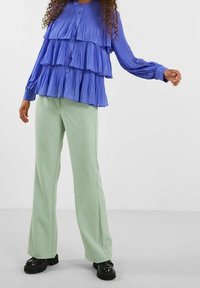 Blusa blu plissettata con tre strati di volants, chiusura con bottoni; abbinata a pantaloni a gamba larga verde chiaro e scarpe platform nere.