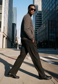 Un homme portant des lunettes de soleil, un pull gris et un pantalon noir marche avec assurance à travers une rue de la ville bordée de hauts immeubles en verre en plein jour.