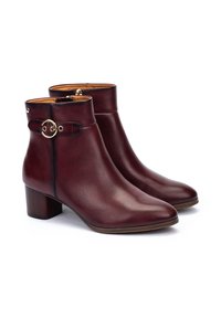 Botas de tobillo de cuero burdeos con punta redonda, tacón cuadrado, acentuación de hebilla circular dorada y detalle de cremallera. Textura suave y diseño elegante.