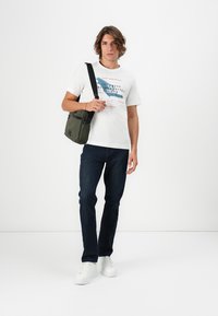 Menns uformelle antrekk med en hvit bomull T-skjorte med grafisk trykk, marineblå denimjeans og en mørkegrønn skulderveske.