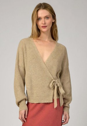 Cardigan en tricot beige avec manches larges, col en V et détail à nouer à la taille, associé à une jupe corail. Texture douce.