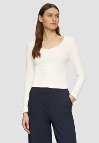 Witte lange mouwen top met een ronde hals en een geribbeld textuur, gedragen met donkere broek met een zijzak. Minimalistisch ontwerp.