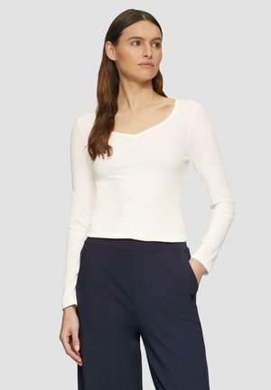 Haut blanc à manches longues avec un décolleté en scoop et une texture côtelée, porté avec un pantalon fonc�é doté une poche latérale. Design minimaliste.