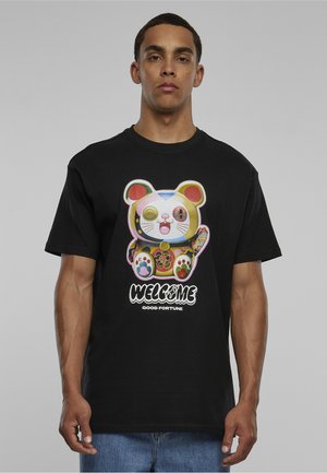 Jeune homme portant un t-shirt noir avec un motif coloré de chat et le texte "WELCOME GOOD FORTUNE" sur le devant, debout devant un fond gris uni.