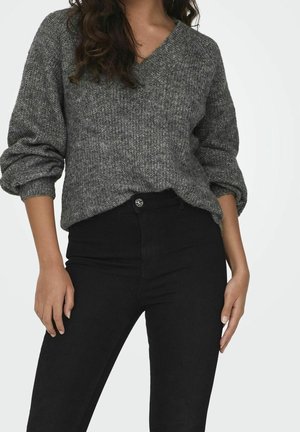 Femme portant un pull en maille gris ample avec les manches retroussées et un pantalon noir ajusté taille haute sur un fond uni.