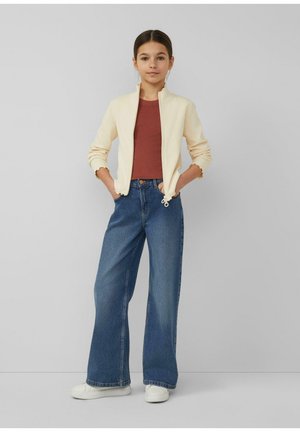 Young girl stands with hands in pockets wearing cream zip jacket, rust red top, wide-leg blue jeans, and white sneakers against plain gray background.