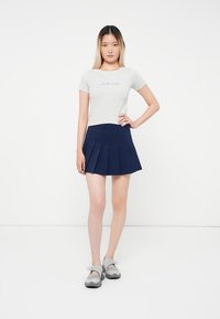 Tânără care stă în picioare, purtând un tricou alb ajustat cu text, o fustă bleumarin plisată și pantofi slip-on gri, pe un fundal alb.