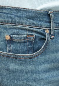 Kék farmernadrág elülső zsebbel, réz szegeccsel és kontrasztos narancssárga varrással. Az anyag sima textúrájú és enyhén kifakult.