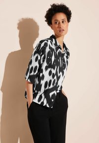 Black and white short-sleeve shirt featuring a bold animal print design, with a collared neckline and a relaxed fit. Paired with black pants.