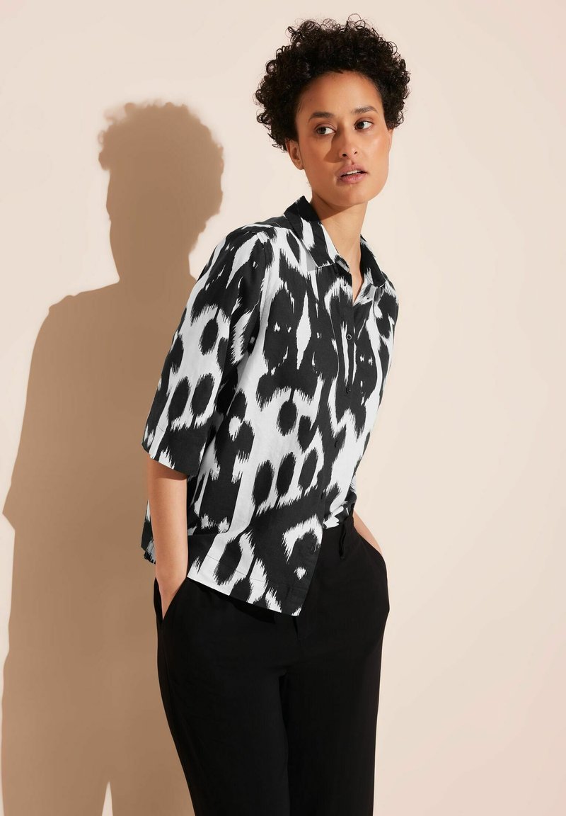 Black and white short-sleeve shirt featuring a bold animal print design, with a collared neckline and a relaxed fit. Paired with black pants.