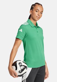 Polo verde con collo, caratterizzato da accenti bianchi a tre strisce sulle spalle. Il modello tiene un pallone da calcio in bianco e nero.