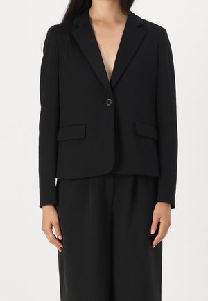 Femme portant un blazer noir à un bouton avec poches rabat, associé à un pantalon noir ample sur un fond uni.