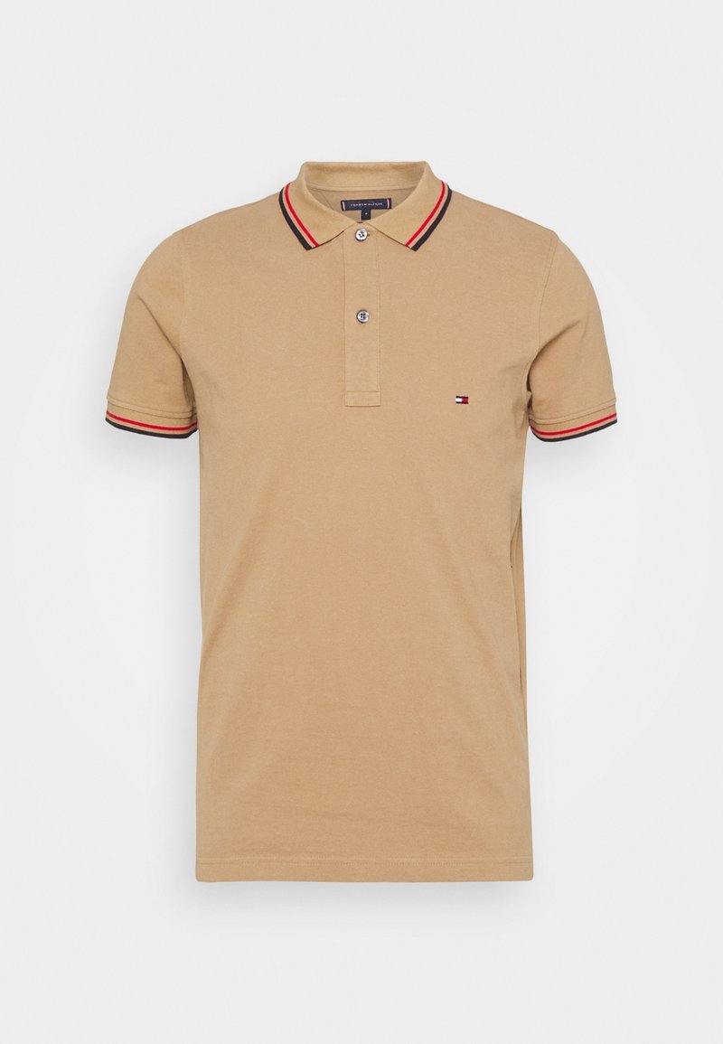 Polo beige à manches courtes avec deux boutons, rayures noires et rouges sur le col et les manches, et petit logo brodé sur le côté gauche de la poitrine.