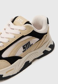 Kraftige sneakers med en beige og svart semsket overdel, meshn paneler, fremhevet logo og en tykk hvit såle med svart mønster.