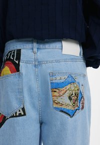 Helles blaues Denim-Jeans mit zwei Gesäßtaschen, die farbenfrohe Grafiken zeigen; eine mit einem brüllenden Gesicht und die andere mit einem mehrfarbigen Muster.