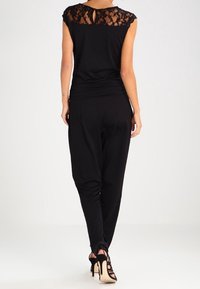 Haut noir avec des détails en dentelle aux épaules, associé à un pantalon noir ajusté. Présente un dos en trou de serrure et une coupe moulante. Talons aiguilles visibles.