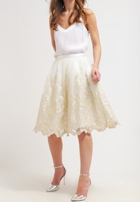 Witte kanten rok met gouden borduursel, volle silhouet, gegolfde zoom, gecombineerd met een witte satijnen top en zilveren hoge hakken.