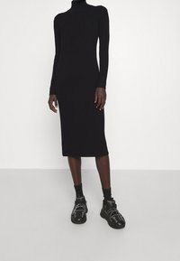 Abito lungo in maglia nera a coste con scollatura alta e linea aderente, abbinato a sneakers nere chunky con dettagli bianchi.