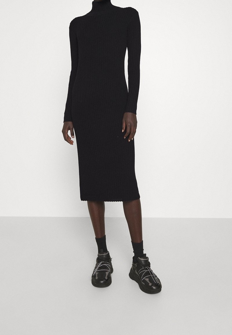 Abito lungo in maglia nera a coste con scollatura alta e linea aderente, abbinato a sneakers nere chunky con dettagli bianchi.