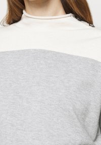 Grå och krämfärgad tröja med hög krage, som har horisontell färgblockering och metalliska knapppdekorer på axeln.