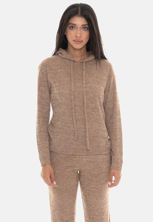 Donna con lunghi capelli scuri che indossa una felpa beige lavorata a maglia con cappuccio e pantaloni abbinati, in piedi davanti a uno sfondo chiaro e uniforme.