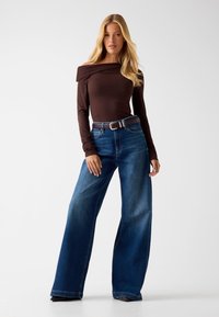 Bruine off-shoulder top met lange mouwen, gecombineerd met losse blauwe high-waisted jeans. Geweven stof met een gladde afwerking en een accent van een riem.