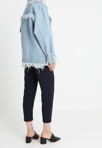 Giacca di jeans azzurro chiaro con bordi sfrangiati, vestibilità ampia e dettagli con bottoni, abbinata a pantaloni navy slim e mule neri con tacco.