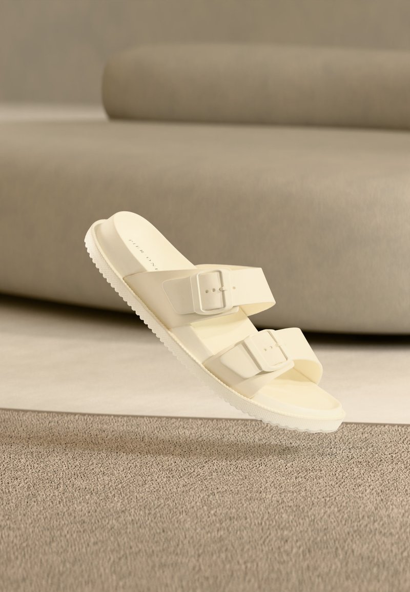 Sandale glissière blanc cassé avec deux brides ajustables à boucle, flottant au-dessus d'un tapis beige et d'un canapé dans un cadre intérieur minimaliste.