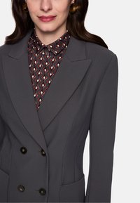 Gray double-breasted blazer with a textured pattern, featuring large black buttons and two front pockets. Underneath, a patterned shirt is visible.