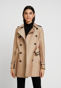 Beige trench coat with double-breasted design, black buttons, and side belt. Features a notched collar and contrasting trim along the cuffs.