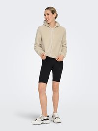 Beige fleece zip-up hoodie with a hood, paired with black biker shorts and white sneakers featuring black and gray accents.