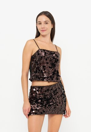 Jeune femme portant un crop top à sequins noir et marron avec fines bretelles et une jupe courte à sequins assortie, debout sur un fond uni.