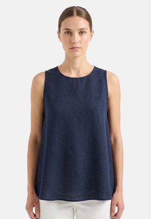 Donna che indossa una canotta in lino blu navy con scollo rotondo e pantaloni bianchi, in piedi davanti a uno sfondo chiaro uniforme.