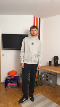 Világosszürke kapucnis pulóver elülső zsebbel, sötét nadrággal és fekete tornacipővel párosítva. Beltéri környezet színes dekorációval és növényekkel.